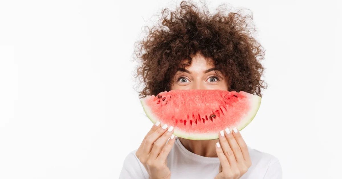Woman holding a watermelon slice in front of her face like a smile.